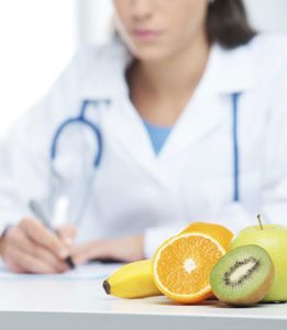 Doctor in a White Lab Coat and Stethoscope Writes on a Notepad at Her White Desk With Cut Fruit Sitting in Front of Her