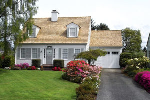 Pretty Home With Tan Roof and White Siding Sits Under a Tree With a Beautifully Landscaped Yard and Asphalt Driveway