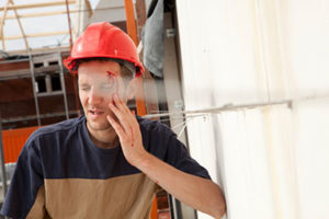 Construction Worker in a Red Hardhat With a Serious Head Wound Grimaces in Pain and Leans Against the Wall of a Construction Site