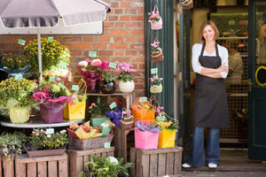 Florist in Black Apron Poses Outside Her Brick Flower Shop, Plants Arranged on Crates Under an Umbrella Outside the Shop
