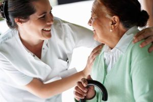 Smiling Nurse Touches the Shoulders of an Elderly Woman in Pearl Earrings and a Green Sweater, Smiling and Holding a Cane